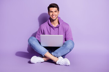 Portrait of attractive cheerful guy sitting lotus pose using laptop free time isolated over purple...