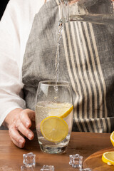 Detail of woman's hand serving tonic in a glass with lemon slices, on wooden table with ice, in vertical