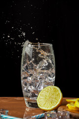 Close-up of glass with tonic and ice with splash, on wooden table with half a lemon and black background, in vertical