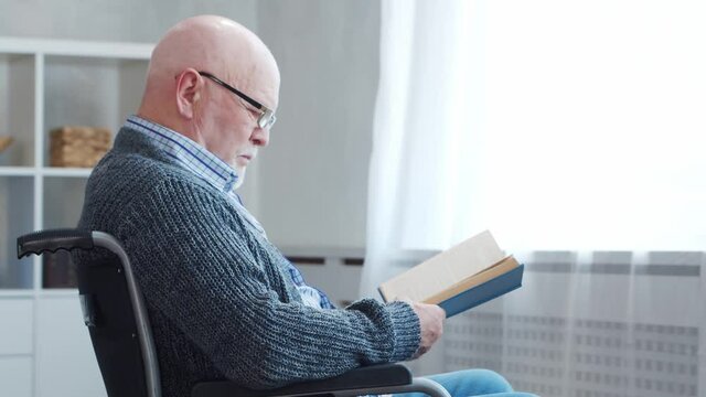 Disabled old man is sitting in a wheelchair at home alone. A handicapped person in a wheelchair is reading the book. Paralysis and disability.
