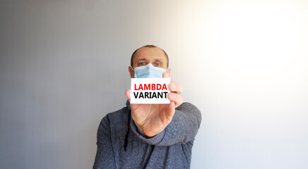 COVID-19 lambda variant symbol. White card with words 'lambda variant'. A young man in a grey wear and medical mask. Sunshine. Beautiful white background. COVID-19 lambda variant coronavirus concept.