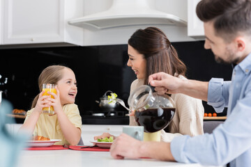 blurred man pouring coffee near smiling daughter and wife talking during breakfast