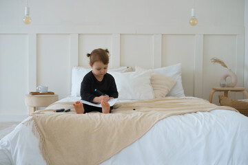 a little girl in black home pajamas is sitting at home on the bed and drawing with felt-tip pens on a piece of paper