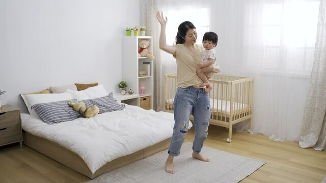 funny asian mother carrying her lovely young kid is moving left and right while dancing barefooted on the carpet in a bright bedroom at home.