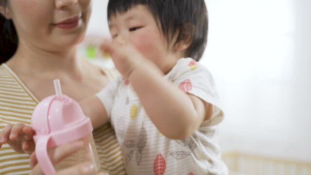 cute thirsty asian baby girl held by her mother is sucking and patting the sippy cup hard while drinking water with closeup shot