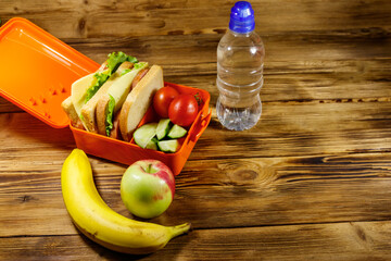 Bottle of water, apple, banana and lunch box with sandwiches and fresh vegetables on a wooden table