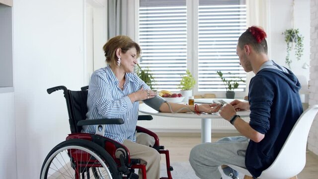 Disabled people in wheelchair sitting at the table indoors at home, measuring blood pressure.