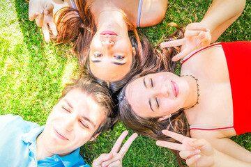 Group of happy teenagers friends relaxing on green grass in meadow and showing peace sign