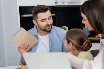 Obraz premium man holding textbook while helping daughter doing homework near wife and blurred laptop