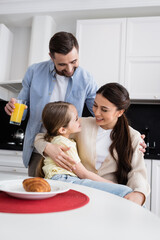 joyful woman embracing daughter near husband with orange juice in kitchen