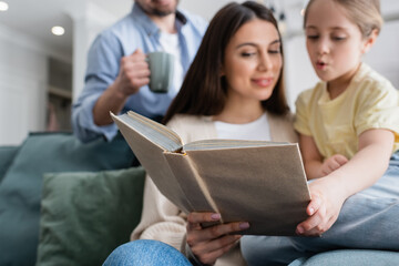 selective focus of book in hand of woman reading to daughter near blurred husband