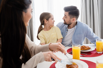 blurred woman cutting croissant near happy daughter and husband looking at each other during breakfast