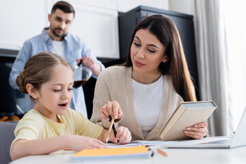woman pointing with pencil while helping daughter doing homework near blurred husband