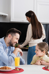 Fototapeta premium happy father and daughter laughing during breakfast in kitchen near blurred mother