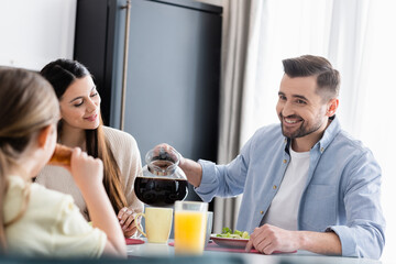 smiling man pouring coffee near wife and blurred daughter during breakfast