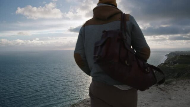 A young male hipster traveler with a bag walks on the path along the cliff in front of beautiful sky and seascape. Traveling alone. Discover new horizons.