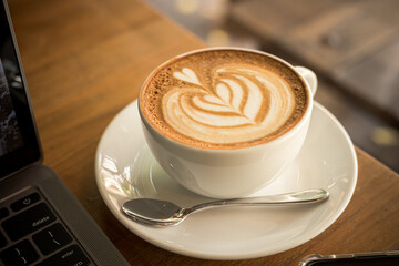 Close-up of Hot coffee latte with latte art milk foam in cup mug and laptop computer and smartphone on wood desk office desk in coffee shop at the cafe,during business work concept
