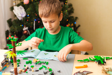 Cute boy collecting block constructor according to instructions sitting by desk