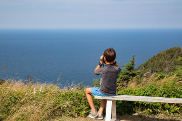 A boy looking at the beautiful ocean of Cape Breton in Nova Scotia through binoculars