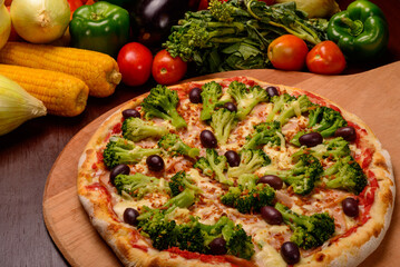 Broccoli pizza with black olives on wooden board and vegetables in the background.