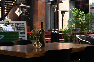 A wooden table on the terrace outside the cafe, in the courtyard, is decorated with a bouquet of daisies in a glass vase and a bottle of wine. Romantic summer vacation of citizens. 