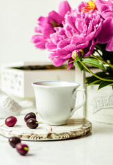 Fototapeta na wymiar Shabby chic tea. A cup of tea on a white wooden tray with a cherry nearby. Peonies are in a white box. Blurred background