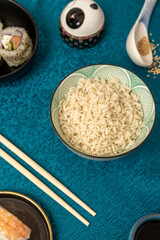 Bowl of white rice with sushi, chopsticks, Chinese blue tablecloth and sesame seeds.