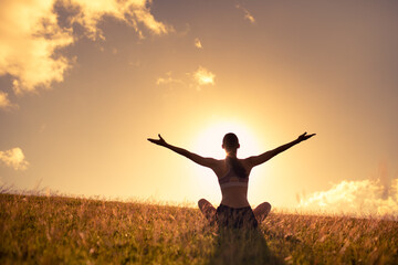 Young happy woman sitting in a grass field lifting her arms up to the sky feeling joy and freedom. 