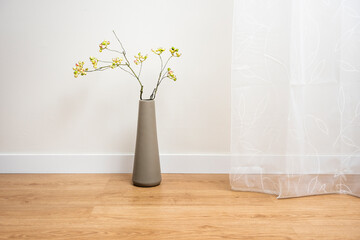 Tall gray vase with decorative branch of green blueberries in room with white curtains and walls