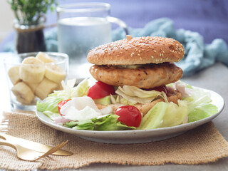 Healthy low calories grilled chicken breast hamburger with high nutrition and protein with fresh green vegetables in a plate such as iceberg, cucumbers and cherry tomatoes. Sliced banana in background