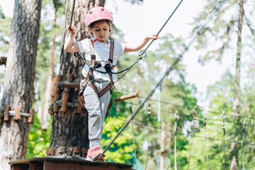 Little girl walks on a rope, hands holding a rope. A child in a rope park passes obstacles
