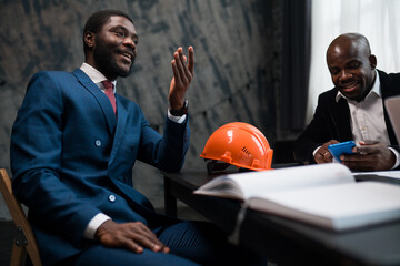 Handsome african american businessman sitting at table negotiating with partner and with construction helmet