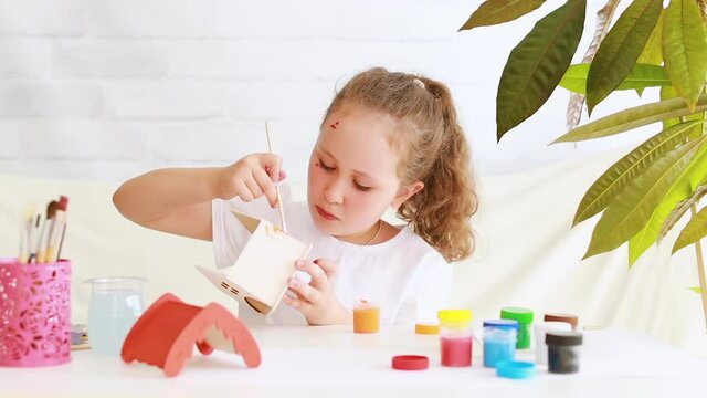 A little girl of preschool age is a creative talented child sitting alone at a table at home, coloring a homemade toy house with colored liquid paints from bottles, studying, doing homework