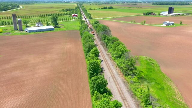 Freight train rolling down tracks, between farm fields on sunny summer day, dynamic moving aerial view.