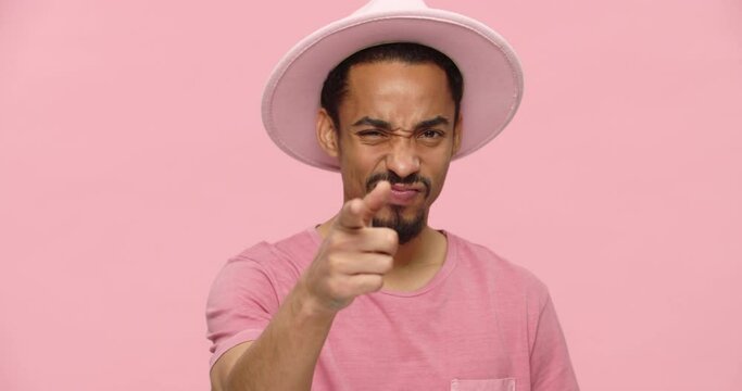 Young displeased black man in pink hat and tshirt pointing with finger to camera and showing killing gesture isolated