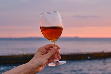 A glass of red wine in the hands of a girl in the rays of the sunset on the seashore. Alcoholic beverage tasting. Beautiful sunset with wine on the beach. Romantic evening aperitif. Glass closeup