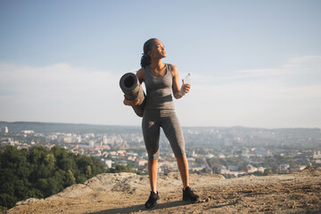 Full length portrait of beautiful african woman in sport clothes standing on high hill with bottle of water and yoga mat in hands. Concept of people, motivation and outdoors activity.