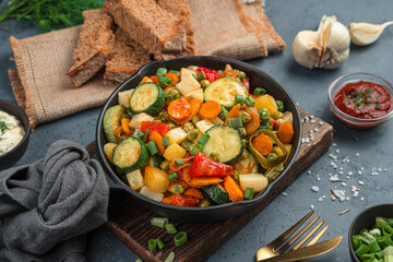 Vegetable stew in a frying pan on a gray background with bread and garlic.
