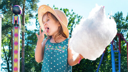 Cute little girl eating a huge piece of cotton candy in an amusement park on a summer day. Happy...