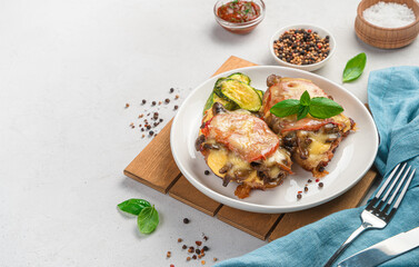 Two pieces of baked chicken breast with mushrooms, tomato and cheese with a side dish of zucchini on a light gray background.