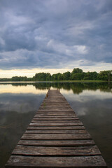 Obraz premium A long wooden footbridge enters the lake. Thick, dark rain clouds above the lake, sunset in the distance