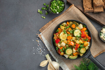 Fried vegetables with fresh herbs in a frying pan on a gray-brown background.