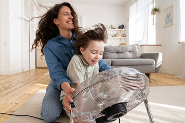 Family bonding: young mom and preschool son laugh sitting together at ventilator working and blowing cool wind in face. Cute child enjoy spending time with parent at home. Parenthood and kids concept