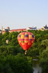 A large multi-colored balloon flies very low over the river and between dense trees. In the background, the buildings of the ancient city