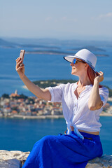 Tourist woman in a white straw hat on beautiful blue Adriatic Sea and cozy island background taking selfie. Blogger women making blog or selfie for social media network. Croatia during summer holiday