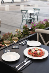 Italian caprese salad with chopped tomatoes, mozzarella, basil, oregano, thassos, onions, radishes, chili peppers and olive oil on a white plate. two glasses of wine rose in a restaurant on the street