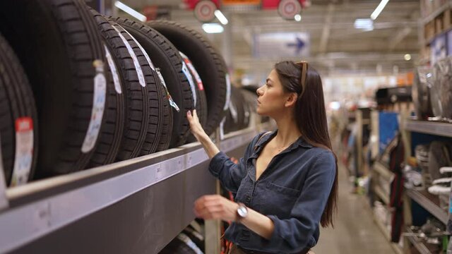 Young woman customer buying new tires while looking at wide assortment represented in department with car goods and accessories in big shopping mall or hypermarket, female in auto shop