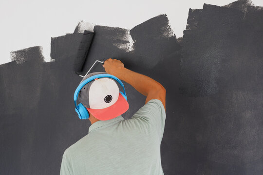 Man Painting A Wall In Black, And Listening Music In Blue Wireless Headphones. Back View.