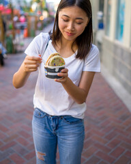 Happy Young Asian Woman Wearing Distressed Jeans and T-Shirt Eating an Ice Cream Sandwich in a Downtown Urban Area