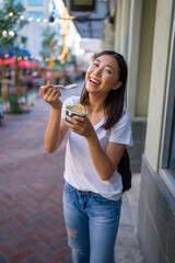 Happy Young Asian Woman Wearing Distressed Jeans and T-Shirt Eating an Ice Cream Sandwich in a Downtown Urban Area